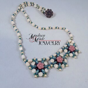 May include: A white and green beaded necklace with three pink flower accents. The necklace has a clasp closure.
