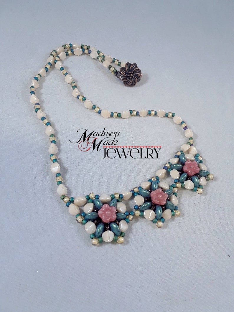 May include: A white and green beaded necklace with three pink flower accents. The necklace has a clasp closure.