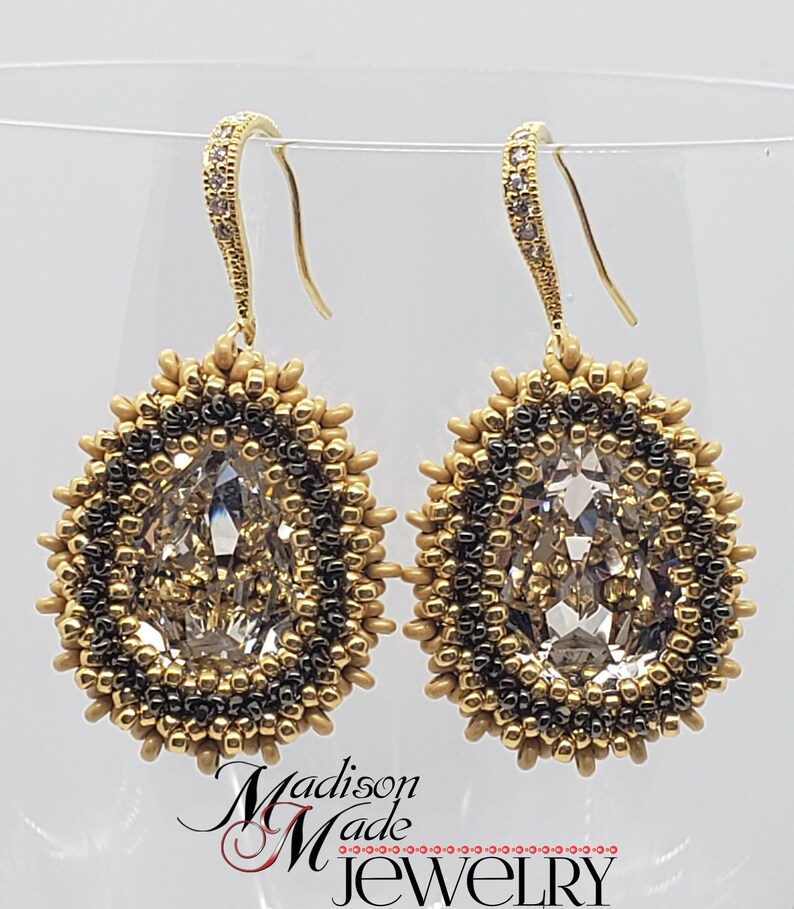 May include: A pair of gold dangle earrings with a clear crystal teardrop stone surrounded by gold and black beads. The earrings have a gold hook closure.