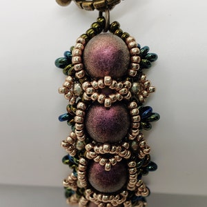 May include: A beaded bracelet with a toggle clasp. The beads are a mix of gold, green, and iridescent purple. The bracelet is labeled "Wine".
