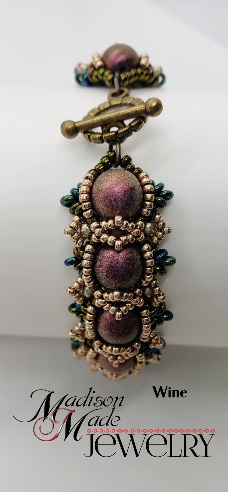 May include: A beaded bracelet with a toggle clasp. The beads are a mix of gold, green, and iridescent purple. The bracelet is labeled "Wine".