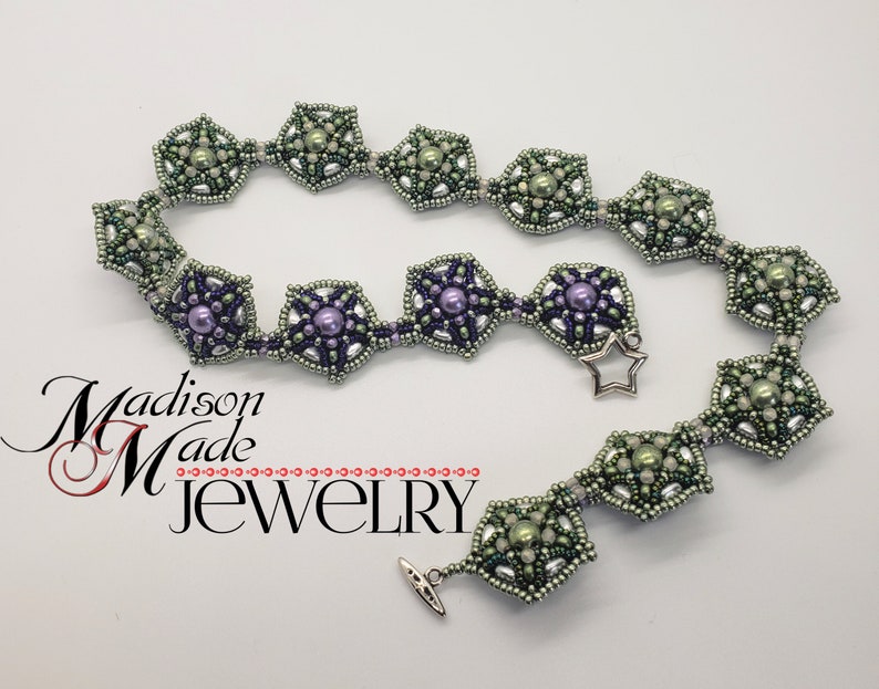 May include: A silver beaded necklace with green and purple beads arranged in a starburst pattern. The necklace has a star-shaped clasp and the text "Madison Made Jewelry" is visible in the lower left corner of the image.