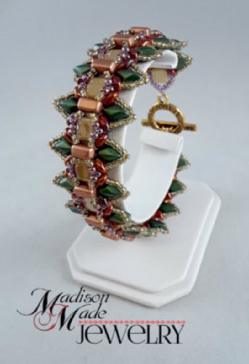 May include: A beaded bracelet with a geometric design. The bracelet features green, copper, and purple beads. It is displayed on a white stand. The bracelet has a gold-colored clasp. The text "Madison Made Jewelry" is at the bottom.