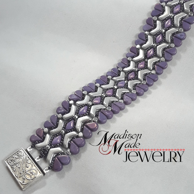 May include: A silver and purple beaded bracelet with a chevron pattern. The bracelet has a silver clasp with an ornate design. The bracelet is made by Madison Made Jewelry.