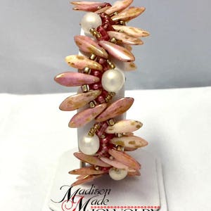 May include: A pink and gold beaded bracelet with white pearls. The bracelet is on a white display stand with the text "Madison Made Jewelry".