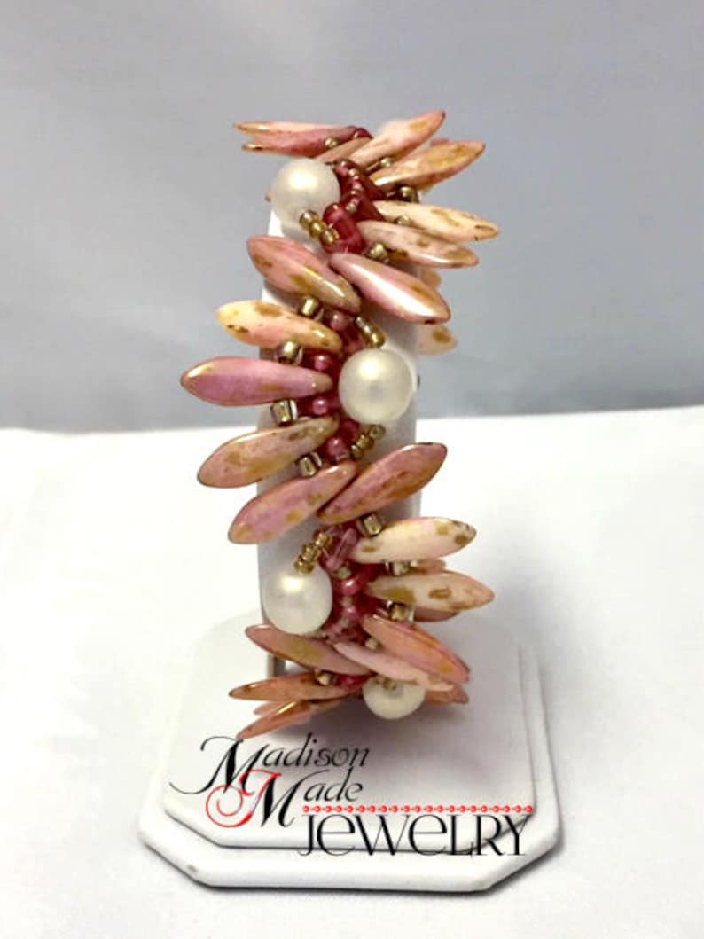 May include: A pink and gold beaded bracelet with white pearls. The bracelet is on a white display stand with the text "Madison Made Jewelry".