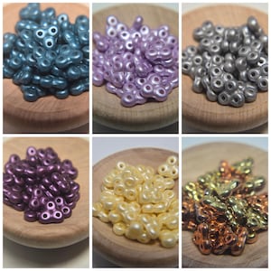 Könnte beinhalten: Sechs kleine Holzschalen, gefüllt mit dekorativen Perlen in verschiedenen Farben. Farben sind Türkis, Lavendel, Silber, Dunkelviolett, Hellgelb und Kupfer. Die Perlen haben ein einzigartiges dreiblättriges Design mit einem Loch in der Mitte.