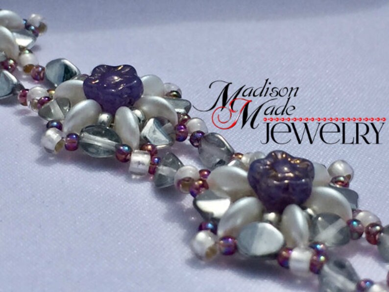 May include: A close-up of a beaded bracelet with white and purple beads. The bracelet features a flower design with a purple center. The text "Madison Made Jewelry" is visible in the background.