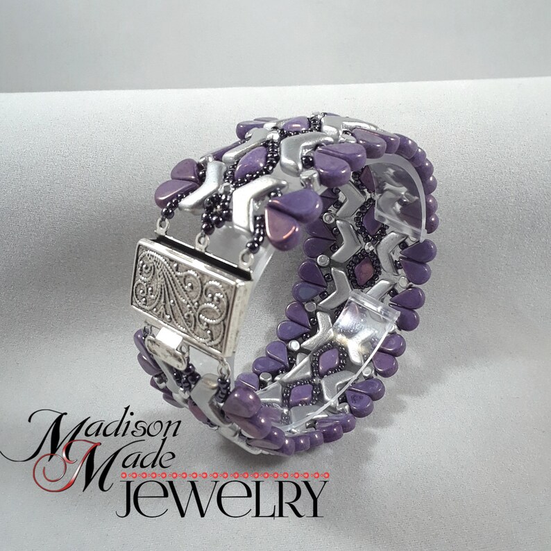 May include: A purple and silver beaded bracelet with a silver clasp. The bracelet is made with a geometric pattern and has a silver clasp with a floral design.
