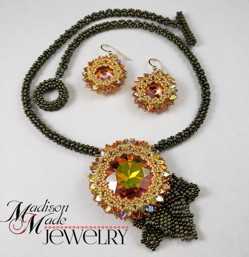 May include: A gold and brown beaded necklace with a large, faceted, orange crystal pendant and matching earrings. The necklace has a tassel-like fringe at the bottom. The text "Madison Made Jewelry" is visible at the bottom of the image.