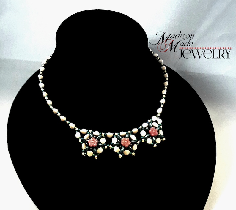 May include: A white and green beaded necklace with pink flower accents. The necklace is displayed on a black jewelry stand. The text "Madison Made JEWELRY" is visible in the upper right corner of the image.