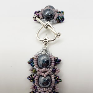 May include: A silver heart-shaped clasp connects to a beaded bracelet with three dark gray hematite beads surrounded by iridescent and pink beads. The bracelet is labeled "Hematite".
