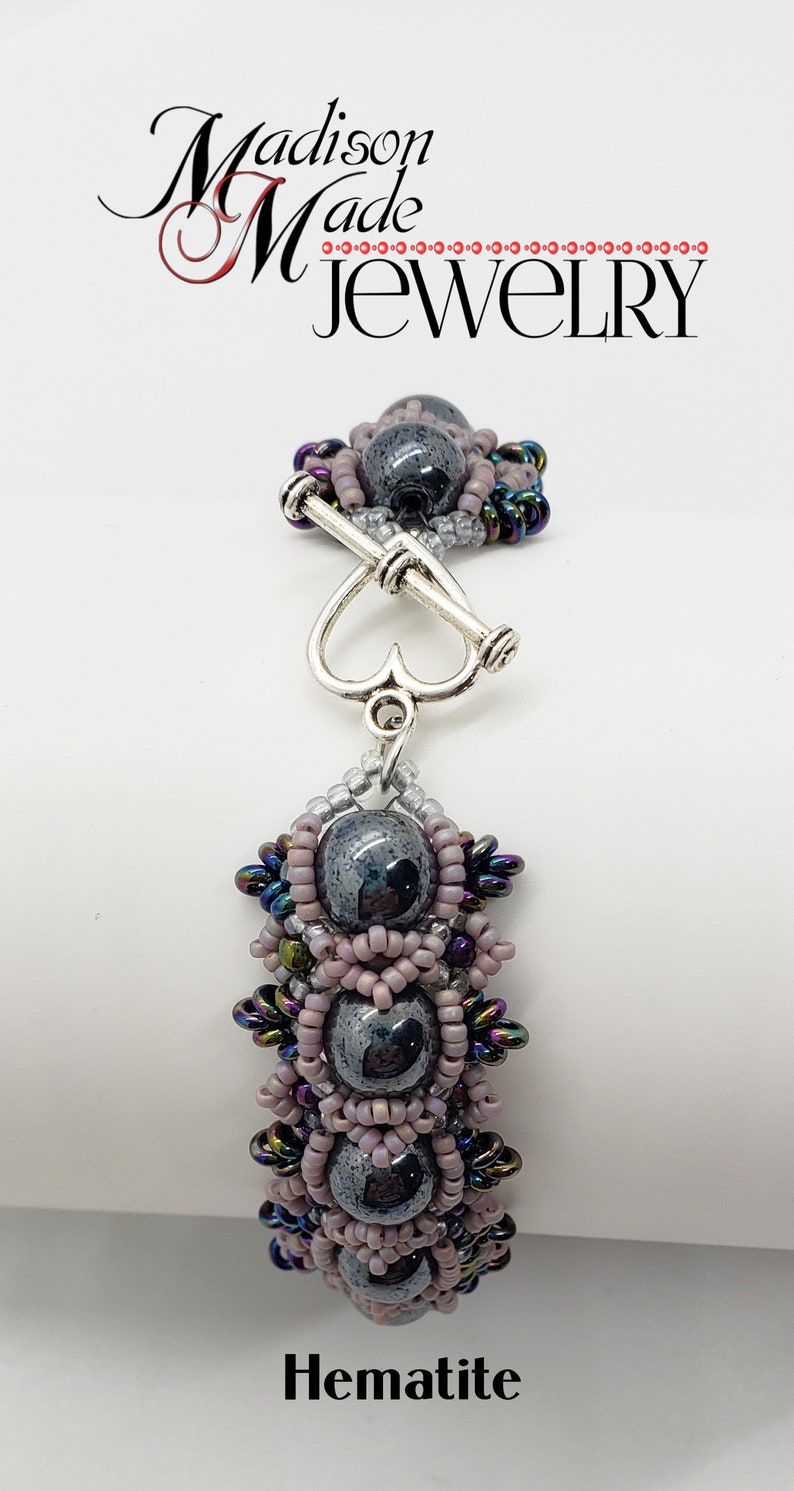 May include: A silver heart-shaped clasp connects to a beaded bracelet with three dark gray hematite beads surrounded by iridescent and pink beads. The bracelet is labeled "Hematite".
