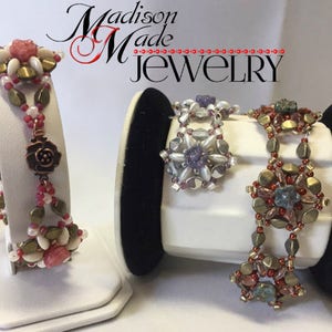 May include: Two beaded bracelets on display stands. The bracelet on the left is made with pink, white, and gold beads. The bracelet on the right is made with blue, white, and gold beads. The bracelets are labeled "Madison Made Jewelry".