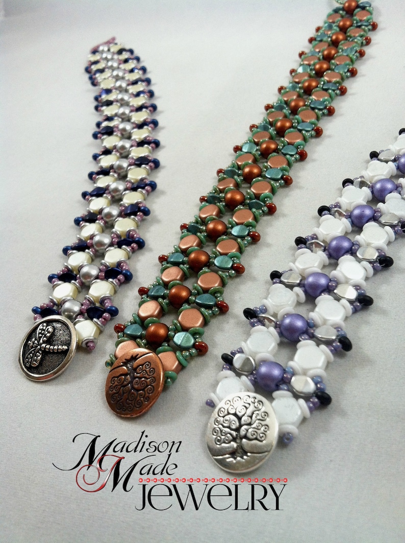 Op de afbeelding: Drie kralenarmbanden met verschillende kleurschema's en decoratieve knoopsluitingen. De armbanden zijn gemaakt van een mix van witte, paarse, blauwe, groene en koperen kralen. De knopen tonen een libel, een boom en een boom met een zilveren afwerking.