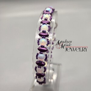 May include: A purple and white beaded bracelet with iridescent beads. The bracelet is on a clear display stand. The text "Madison Made Jewelry" is visible in the background.