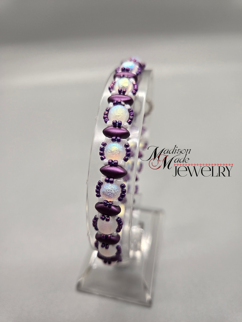 May include: A purple and white beaded bracelet with iridescent beads. The bracelet is on a clear display stand. The text "Madison Made Jewelry" is visible in the background.