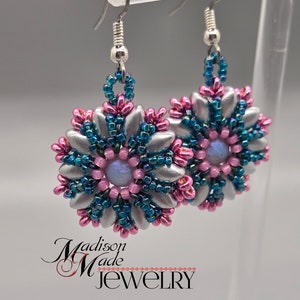 Może przedstawiać: Para ręcznie robionych kolczyków z koralików. Każdy kolczyk ma srebrny wzór płatków, zaakcentowany turkusowymi i różowymi koralikami. Środek każdego kolczyka ma jasnofioletowy koralik. Widoczny jest tekst "Madison Made Jewelry".
