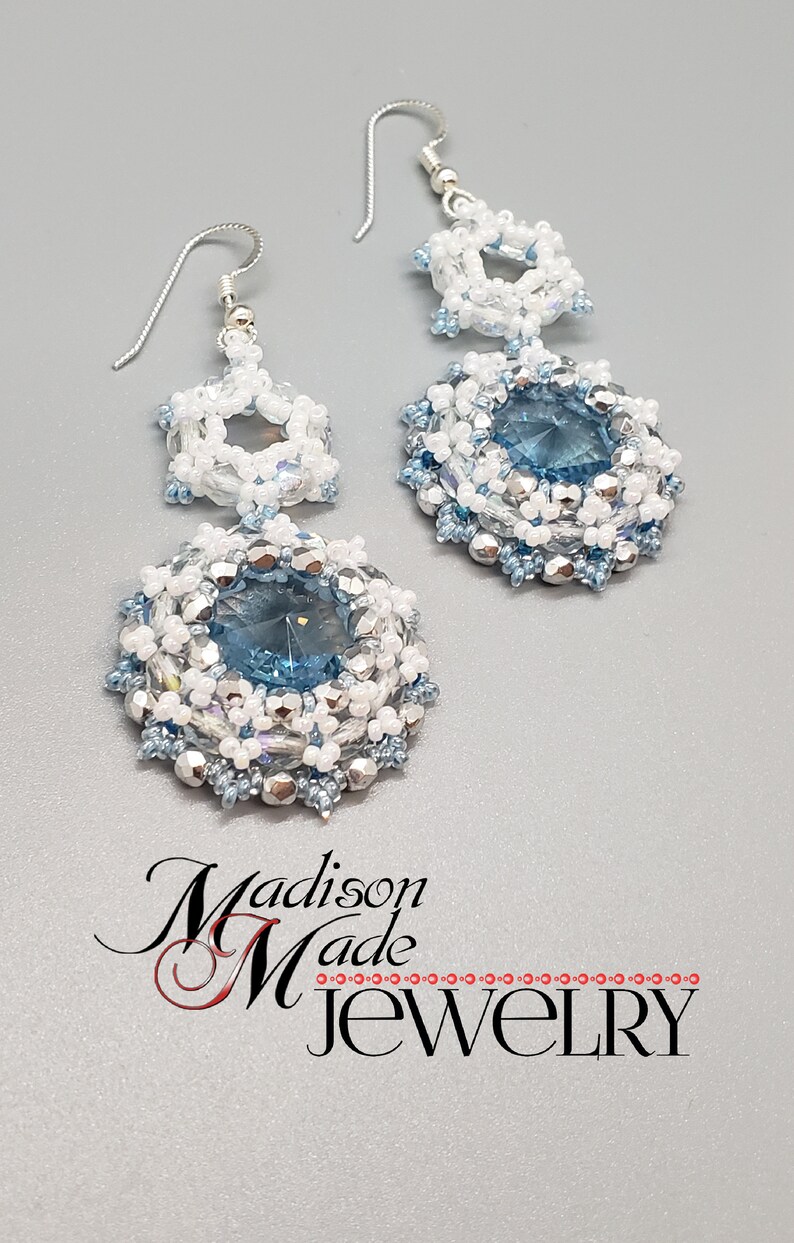 May include: A pair of silver earrings with blue and white crystal beads. The earrings feature a larger round bead with a blue crystal in the center, surrounded by smaller white and silver beads. The earrings are attached to silver hooks.