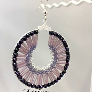 May include: A pair of silver hoop earrings with a beaded design. The beads are in shades of purple and black, creating a gradient effect. The earrings are handmade and feature a unique, circular design.