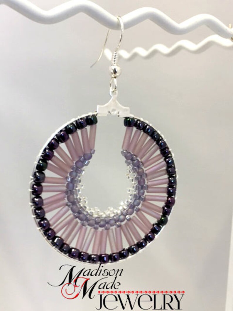 May include: A pair of silver hoop earrings with a beaded design. The beads are in shades of purple and black, creating a gradient effect. The earrings are handmade and feature a unique, circular design.