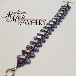 Könnte beinhalten: Ein Perlenarmband mit Knebelverschluss. Das Armband hat ein Muster aus silberfarbenen rechteckigen Perlen, durchsetzt mit dunkelblauen gesprenkelten Perlen und weinroten runden Perlen. Der Text "Madison Made Jewelry" befindet sich in der oberen linken Ecke.