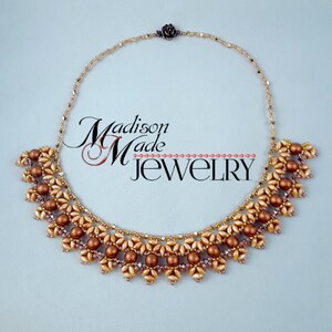 May include: A gold and brown beaded necklace with a flower design. The necklace is made with small seed beads and larger round beads. The necklace is on a light blue background. Madison Made Jewelry.