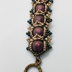May include: A beaded bracelet with a bronze clasp. The beads are a mix of gold, silver, and dark purple. The bracelet is labeled "Wine".