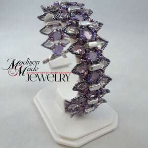 May include: A beaded bracelet with a floral design. The bracelet features a combination of purple, silver, and clear beads. The bracelet is displayed on a white stand. The text "Madison Made Jewelry" is visible.