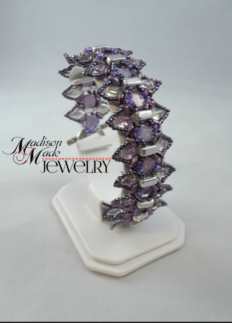 May include: A beaded bracelet with a floral design. The bracelet features a combination of purple, silver, and clear beads. The bracelet is displayed on a white stand. The text "Madison Made Jewelry" is visible.