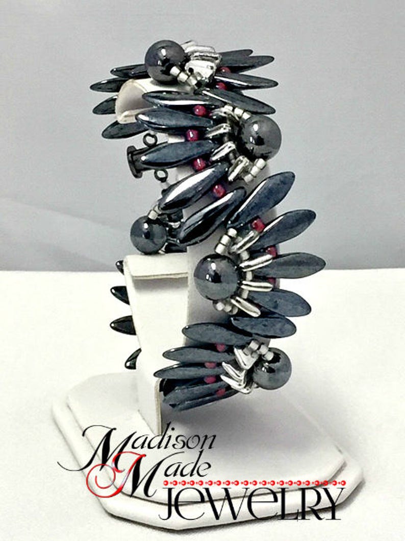 May include: A black and silver bracelet with red beads. The bracelet is made of small, pointed beads and is displayed on a white stand. The stand has the text "Madison Made Jewelry" on it.