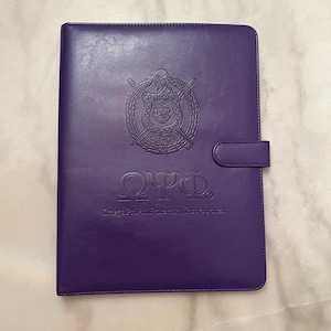 May include: A purple leather binder with a gold embossed crest and the text "ΩΨΦ. Omega Psi Phi Fraternity, Incorporated."