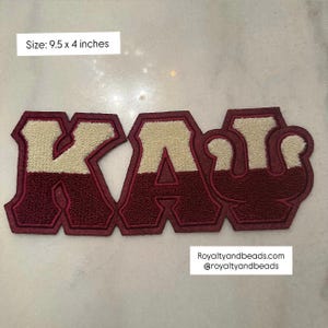 May include: A maroon and cream-colored embroidered patch with the letters KAU. The patch is 9.5 x 4 inches. The letters are outlined in maroon with a cream-colored center. The patch has a textured appearance.