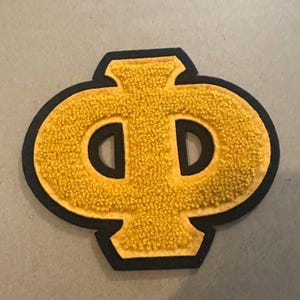 May include: A yellow felt letterman patch with a black border. The patch features a stylized letter "Φ" in a textured, golden-yellow color. The design is a classic varsity style, suitable for jackets or bags.