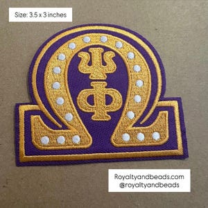 May include: An embroidered patch in purple and gold featuring the Greek letter Omega, with the letters Psi and Phi centered inside. The design includes white circular accents along the Omega shape. The patch measures 3.5 x 3 inches.