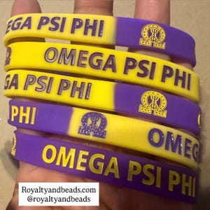 May include: A stack of yellow and purple rubber bracelets. Each bracelet has the text "OMEGA PSI PHI" and a Greek symbol. The bracelets are two-toned, with a yellow and purple color scheme.