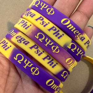 May include: A stack of purple and yellow rubber bracelets. The bracelets have the Greek letters Omega, Psi, and Phi, along with the words "Omega Psi Phi" in gold lettering. The bracelets are approximately 0.5 inches wide.