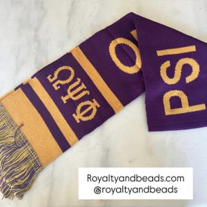 May include: A purple and gold knit scarf with the Greek letters Omega, Psi, and Phi, and the word "Psi" in gold. The scarf has gold fringe at the end and is displayed on a white surface.