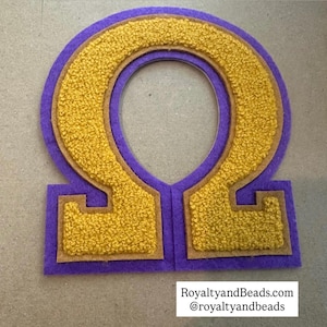May include: A purple and gold felt letter patch in the shape of the Greek letter Omega. The Omega is outlined in gold and filled with textured gold fabric. The patch has a brown border and the text "RoyaltyandBeads.com @royaltyandbeads".