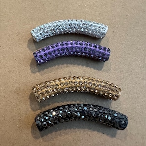 May include: Four curved, rhinestone-covered beads in silver, purple, gold, and black. The beads are arranged in a row, each with a slightly different color and sparkle. The beads are likely for jewelry making.
