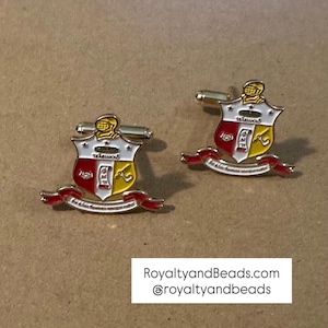 May include: Pair of silver cufflinks with a shield design in red, white, and yellow. The shield features a knight's helmet and Greek letters. A red banner with text is below the shield. The cufflinks are on a tan surface.