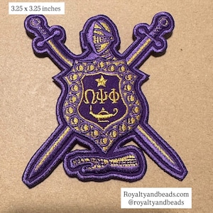 May include: Embroidered purple patch, 3.25 x 3.25 inches, featuring a shield design with a knight's helmet, crossed swords, and Greek letters. The shield has a gold star and a lamp design. The patch has gold accents.