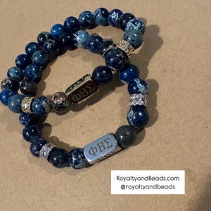 May include: Two beaded bracelets featuring dark blue and speckled beads. Each bracelet has a decorative metal accent with the Greek letters ΦΒΣ and rhinestone spacers. The bracelets are displayed on a neutral surface.