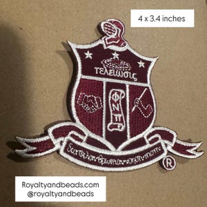 May include: Embroidered maroon and white crest patch, measuring 4 x 3.4 inches. The patch features a shield design with Greek lettering and symbols, including a handshake and a scroll. A banner at the bottom has more text. The patch is bordered with white stitching.