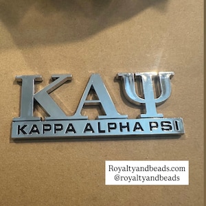 May include: Silver metal emblem featuring the Greek letters K A Ψ, with the words KAPPA ALPHA PSI below. The emblem is set against a brown background. The text "Royaltyandbeads.com @royaltyandbeads" is visible.