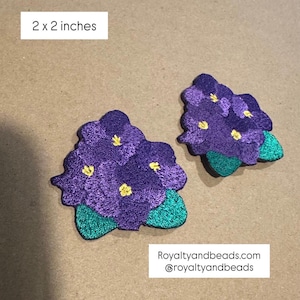 May include: Two embroidered violet flower patches, each measuring 2 x 2 inches. The flowers are a deep purple with yellow centers and green leaves. The patches are likely for decorative purposes.