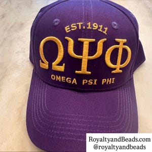 May include: A purple baseball cap with gold embroidered text. The text includes "EST.1911", and the Greek letters Omega, Psi, and Phi, along with "OMEGA PSI PHI". The cap has a curved brim and a structured crown.
