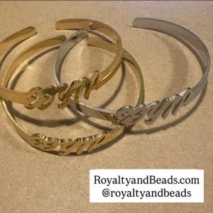 May include: Gold and silver-toned metal cuff bracelets. Each bracelet features a name in a cursive font. The bracelets are displayed on a neutral surface. The text "RoyaltyandBeads.com @royaltyandbeads" is visible at the bottom.