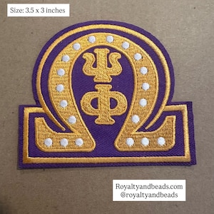 May include: A purple and gold embroidered patch, measuring 3.5 x 3 inches. The design features a gold emblem with white dots, set against a purple background. The patch has a rectangular shape with a curved top.