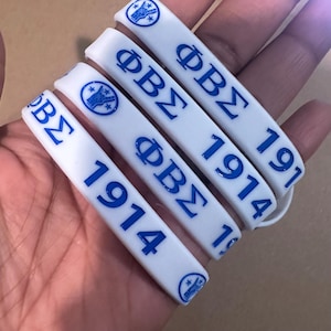 May include: Four white silicone bracelets with blue lettering. Each bracelet has the text "ΦΒΣ" and the year "1914". The bracelets have a small blue logo with a hand and three stars.